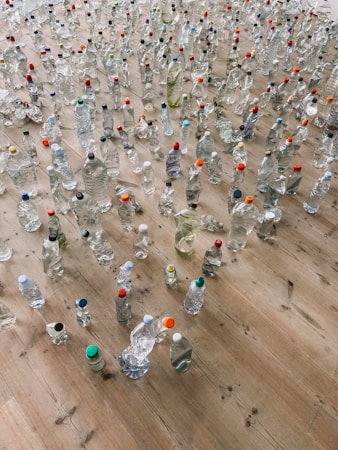 A large collection of plastic bottles of various shapes and sizes spread across a wooden floor. Some bottles are clear while others contain liquids of different colors. Many have colorful caps in shades like red, blue, green, and orange.