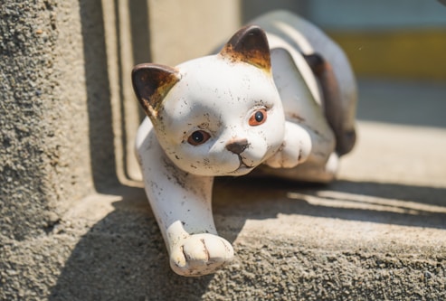 A ceramic or stone sculpture of a cat is positioned on a textured concrete surface, with the cat in a playful crouching posture. The sculpture has a smooth white finish with hints of brown on the ears. The scene is well-lit, casting soft shadows that add to the realism of the sculpture.