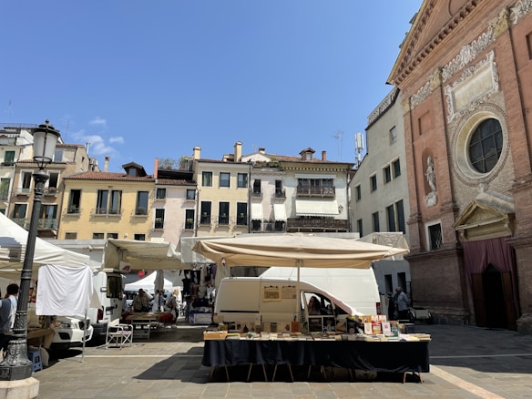A lively outdoor market is set up in a quaint European square. Stalls with white canopies are arranged around a stone plaza, with vendors selling items like books. Behind the market, a picturesque row of old buildings with colorful facades and wrought-iron balconies provide a charming backdrop. To the right, an ornate church with detailed carvings and large circular window catches the eye. The sky is clear and bright blue, creating a vibrant atmosphere.