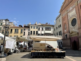 A lively outdoor market is set up in a quaint European square. Stalls with white canopies are arranged around a stone plaza, with vendors selling items like books. Behind the market, a picturesque row of old buildings with colorful facades and wrought-iron balconies provide a charming backdrop. To the right, an ornate church with detailed carvings and large circular window catches the eye. The sky is clear and bright blue, creating a vibrant atmosphere.