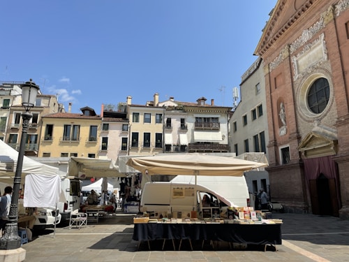 A lively outdoor market is set up in a quaint European square. Stalls with white canopies are arranged around a stone plaza, with vendors selling items like books. Behind the market, a picturesque row of old buildings with colorful facades and wrought-iron balconies provide a charming backdrop. To the right, an ornate church with detailed carvings and large circular window catches the eye. The sky is clear and bright blue, creating a vibrant atmosphere.