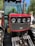 A close-up view of the front grille and headlights of a red tractor with robust metal bars and large tires. The background includes blurred greenery and a person standing nearby.