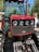 A close-up view of the front grille and headlights of a red tractor with robust metal bars and large tires. The background includes blurred greenery and a person standing nearby.