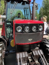 A close-up view of the front grille and headlights of a red tractor with robust metal bars and large tires. The background includes blurred greenery and a person standing nearby.