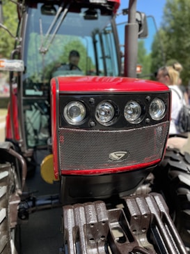 A close-up view of the front grille and headlights of a red tractor with robust metal bars and large tires. The background includes blurred greenery and a person standing nearby.