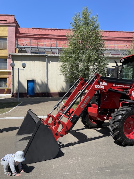 A red tractor with a large front loader is parked on a paved area beside a building. A child wearing a sun hat and striped clothing crouches near the loader's bucket. The background shows a yellow and pink building, with a tree providing some shade.