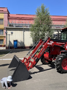 A red tractor with a large front loader is parked on a paved area beside a building. A child wearing a sun hat and striped clothing crouches near the loader's bucket. The background shows a yellow and pink building, with a tree providing some shade.