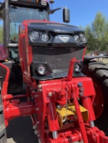 A close-up view of the front part of a red and black tractor, focusing on the headlights and grill. The tractor is parked outdoors, with clear blue sky and trees visible in the background.