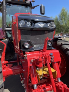 A close-up view of the front part of a red and black tractor, focusing on the headlights and grill. The tractor is parked outdoors, with clear blue sky and trees visible in the background.
