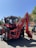 A red tractor with a front loader attachment is parked on a street. There are people around, including one individual working near the rear tire and another adjusting something near the cabin. The tractor has a marked number 20 and is labeled as a Belarus brand. The background includes tall trees and a red building under a clear blue sky.