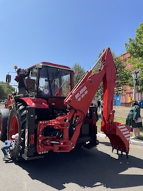 A red tractor with a front loader attachment is parked on a street. There are people around, including one individual working near the rear tire and another adjusting something near the cabin. The tractor has a marked number 20 and is labeled as a Belarus brand. The background includes tall trees and a red building under a clear blue sky.