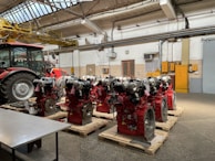 Several industrial engines with red components are neatly arranged on wooden pallets inside a workshop with a tractor nearby. The environment features concrete floors, a high ceiling with metal beams, and overhead lighting. Yellow pipes and equipment are visible on the walls, adding to the industrial atmosphere.