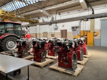 Several industrial engines with red components are neatly arranged on wooden pallets inside a workshop with a tractor nearby. The environment features concrete floors, a high ceiling with metal beams, and overhead lighting. Yellow pipes and equipment are visible on the walls, adding to the industrial atmosphere.