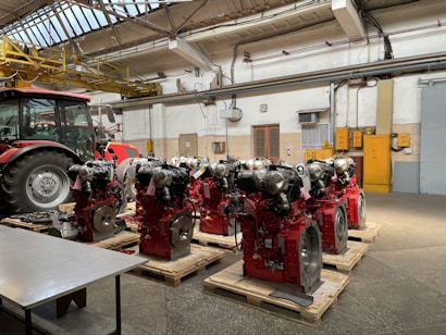 Several industrial engines with red components are neatly arranged on wooden pallets inside a workshop with a tractor nearby. The environment features concrete floors, a high ceiling with metal beams, and overhead lighting. Yellow pipes and equipment are visible on the walls, adding to the industrial atmosphere.