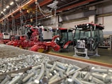 An industrial assembly area featuring several red agricultural machines in various stages of assembly. The foreground shows a bin filled with metal bolts. Overhead, there are cranes and hooks, suggesting a manufacturing environment. The machinery has visible engines and glass cabins.