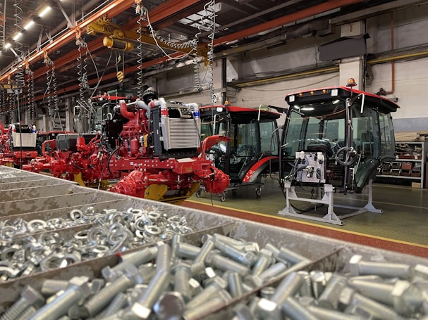 An industrial assembly area featuring several red agricultural machines in various stages of assembly. The foreground shows a bin filled with metal bolts. Overhead, there are cranes and hooks, suggesting a manufacturing environment. The machinery has visible engines and glass cabins.