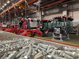An industrial assembly area featuring several red agricultural machines in various stages of assembly. The foreground shows a bin filled with metal bolts. Overhead, there are cranes and hooks, suggesting a manufacturing environment. The machinery has visible engines and glass cabins.