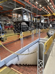 A spacious industrial facility with multiple tractor cabs lined up in sequence. The setting includes bright overhead lighting with orange and yellow industrial pipework on the ceiling. Safety barriers surround a stairway leading downward, and the ground is painted a reddish-brown color.