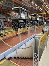 A spacious industrial facility with multiple tractor cabs lined up in sequence. The setting includes bright overhead lighting with orange and yellow industrial pipework on the ceiling. Safety barriers surround a stairway leading downward, and the ground is painted a reddish-brown color.