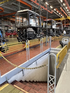 A spacious industrial facility with multiple tractor cabs lined up in sequence. The setting includes bright overhead lighting with orange and yellow industrial pipework on the ceiling. Safety barriers surround a stairway leading downward, and the ground is painted a reddish-brown color.