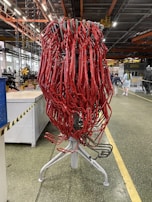 A large cluster of red tubes intricately intertwined and mounted on a metallic stand is situated in an industrial setting. The background features a typical factory environment with visible machinery, overhead piping, and industrial lighting. A few people are seen walking in the distance.