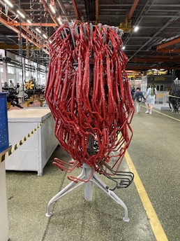 A large cluster of red tubes intricately intertwined and mounted on a metallic stand is situated in an industrial setting. The background features a typical factory environment with visible machinery, overhead piping, and industrial lighting. A few people are seen walking in the distance.