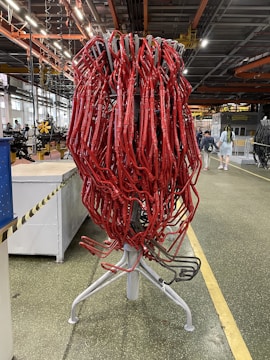 A large cluster of red tubes intricately intertwined and mounted on a metallic stand is situated in an industrial setting. The background features a typical factory environment with visible machinery, overhead piping, and industrial lighting. A few people are seen walking in the distance.