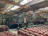 An industrial factory setting with machinery and equipment. The foreground contains numerous red plastic containers arranged in rows. Overhead, metal beams support the roof with large windows allowing natural light to illuminate the space. Pipes and control panels are visible, indicating a manufacturing operation.