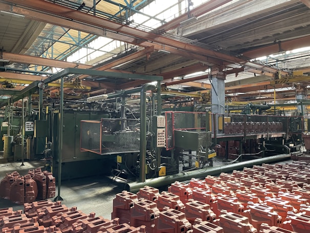 An industrial factory setting with machinery and equipment. The foreground contains numerous red plastic containers arranged in rows. Overhead, metal beams support the roof with large windows allowing natural light to illuminate the space. Pipes and control panels are visible, indicating a manufacturing operation.