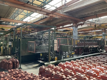 An industrial factory setting with machinery and equipment. The foreground contains numerous red plastic containers arranged in rows. Overhead, metal beams support the roof with large windows allowing natural light to illuminate the space. Pipes and control panels are visible, indicating a manufacturing operation.