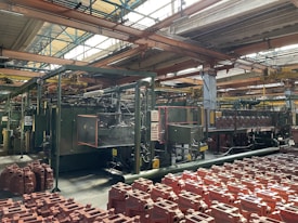 An industrial factory setting with machinery and equipment. The foreground contains numerous red plastic containers arranged in rows. Overhead, metal beams support the roof with large windows allowing natural light to illuminate the space. Pipes and control panels are visible, indicating a manufacturing operation.