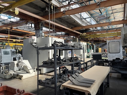 An industrial setting with various large machines and equipment, including a CNC machine and other manufacturing tools. There are shelves stocked with metal parts and components, and the space is illuminated by natural light streaming in through the ceiling windows. The area appears organized with clear sections for different types of machinery.