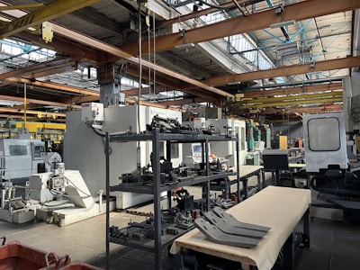 An industrial setting with various large machines and equipment, including a CNC machine and other manufacturing tools. There are shelves stocked with metal parts and components, and the space is illuminated by natural light streaming in through the ceiling windows. The area appears organized with clear sections for different types of machinery.