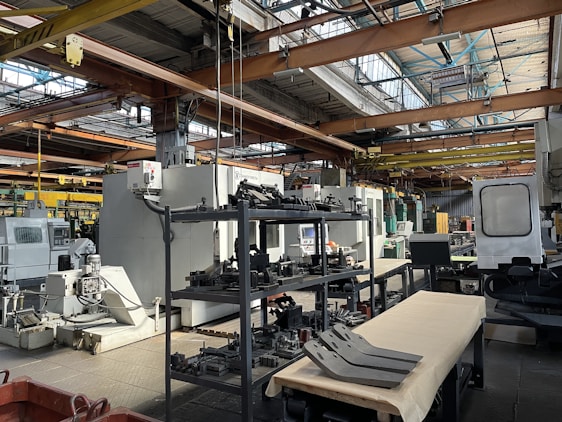An industrial setting with various large machines and equipment, including a CNC machine and other manufacturing tools. There are shelves stocked with metal parts and components, and the space is illuminated by natural light streaming in through the ceiling windows. The area appears organized with clear sections for different types of machinery.
