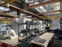 An industrial setting with various large machines and equipment, including a CNC machine and other manufacturing tools. There are shelves stocked with metal parts and components, and the space is illuminated by natural light streaming in through the ceiling windows. The area appears organized with clear sections for different types of machinery.
