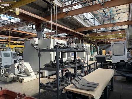 An industrial setting with various large machines and equipment, including a CNC machine and other manufacturing tools. There are shelves stocked with metal parts and components, and the space is illuminated by natural light streaming in through the ceiling windows. The area appears organized with clear sections for different types of machinery.