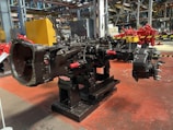 A large, industrial machine component is centrally positioned in a factory setting. The machinery appears to be part of a mechanical assembly line, mounted on a sturdy black stand with a metallic surface. The background includes structural beams and various pieces of industrial equipment, with a focus on mechanical and robust engineering. The floor is painted a rusty red and the walls have industrial elements such as piping and storage areas.