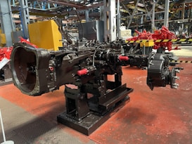 A large, industrial machine component is centrally positioned in a factory setting. The machinery appears to be part of a mechanical assembly line, mounted on a sturdy black stand with a metallic surface. The background includes structural beams and various pieces of industrial equipment, with a focus on mechanical and robust engineering. The floor is painted a rusty red and the walls have industrial elements such as piping and storage areas.