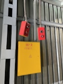 A yellow electrical box with a warning symbol and red lettering is mounted on a corrugated metal wall. Two red control devices with buttons are hanging from gray cables above the box. The environment appears industrial, with structural beams visible.
