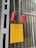A yellow electrical box with a warning symbol and red lettering is mounted on a corrugated metal wall. Two red control devices with buttons are hanging from gray cables above the box. The environment appears industrial, with structural beams visible.