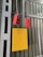 A yellow electrical box with a warning symbol and red lettering is mounted on a corrugated metal wall. Two red control devices with buttons are hanging from gray cables above the box. The environment appears industrial, with structural beams visible.