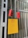 A yellow electrical box with a warning symbol and red lettering is mounted on a corrugated metal wall. Two red control devices with buttons are hanging from gray cables above the box. The environment appears industrial, with structural beams visible.
