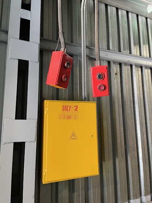 A yellow electrical box with a warning symbol and red lettering is mounted on a corrugated metal wall. Two red control devices with buttons are hanging from gray cables above the box. The environment appears industrial, with structural beams visible.