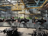 An industrial setting inside a factory with multiple large mechanical components arranged in a row. A complex network of beams, pipes, and metal structures is visible above, indicating a manufacturing or assembly environment. The lighting is dim and utilitarian, focusing on functionality rather than aesthetics.
