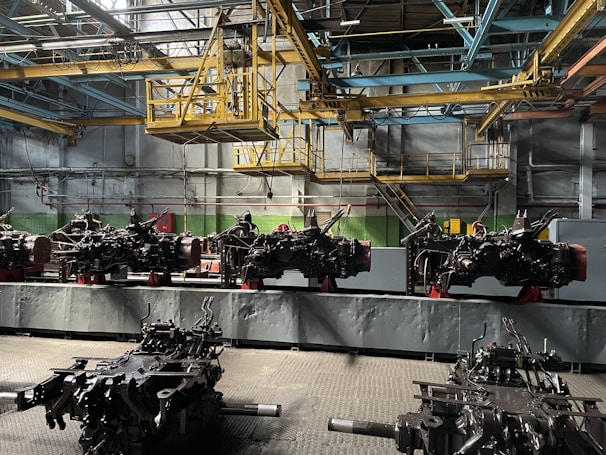 An industrial setting inside a factory with multiple large mechanical components arranged in a row. A complex network of beams, pipes, and metal structures is visible above, indicating a manufacturing or assembly environment. The lighting is dim and utilitarian, focusing on functionality rather than aesthetics.