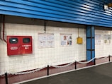 A tiled wall featuring a red fire equipment box and several informational posters. The setting appears to be industrial or public, with blue corrugated metal siding above the wall and a chain barrier in the foreground.
