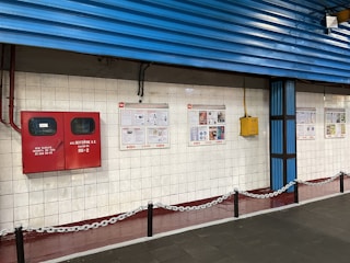 A tiled wall featuring a red fire equipment box and several informational posters. The setting appears to be industrial or public, with blue corrugated metal siding above the wall and a chain barrier in the foreground.