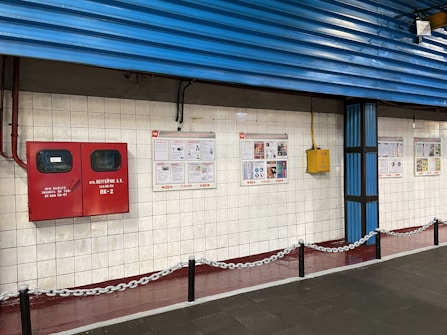 A tiled wall featuring a red fire equipment box and several informational posters. The setting appears to be industrial or public, with blue corrugated metal siding above the wall and a chain barrier in the foreground.