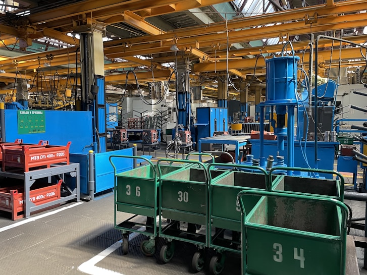 An industrial facility with numerous pieces of machinery, metal carts labeled with numbers, and various colorful equipment. Overhead, yellow girders and cranes are visible, and wires or hoses hang throughout the space.