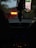 The scene takes place inside a car with a GPS on the dashboard. Dim lighting outside suggests evening or night, complementing the glow of the electronic displays. Hanging from the rear-view mirror are assorted objects.